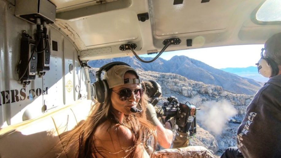 A smiling woman wearing aviators in a helicopter beside a door gunner shooting over the Nevada desert